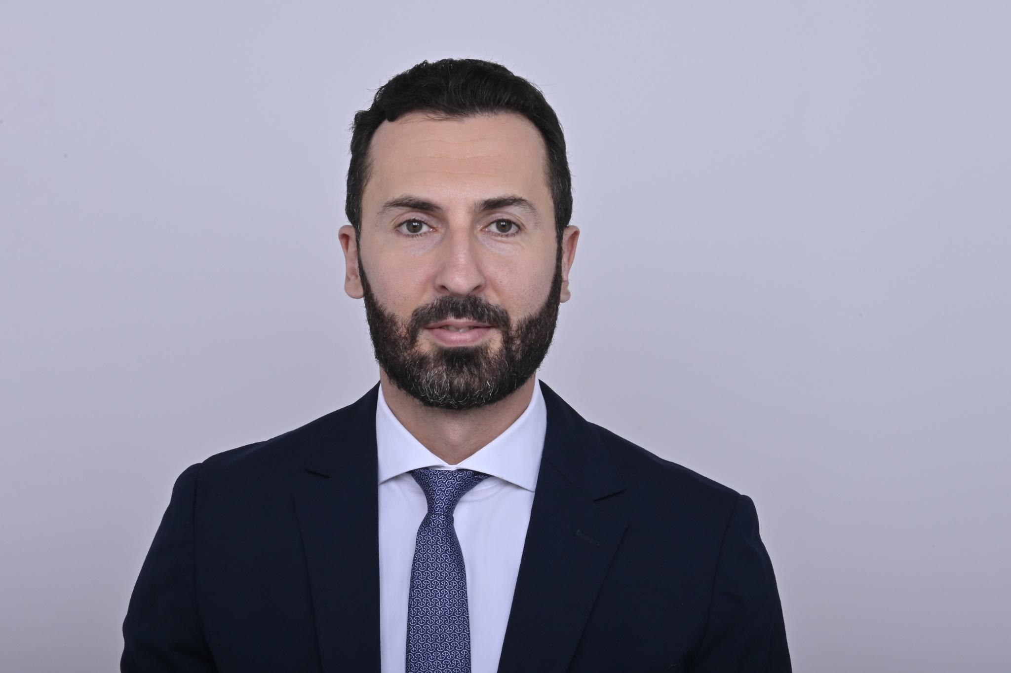 A man with a beard and wearing a suit is looking directly at the camera, set against a plain background.